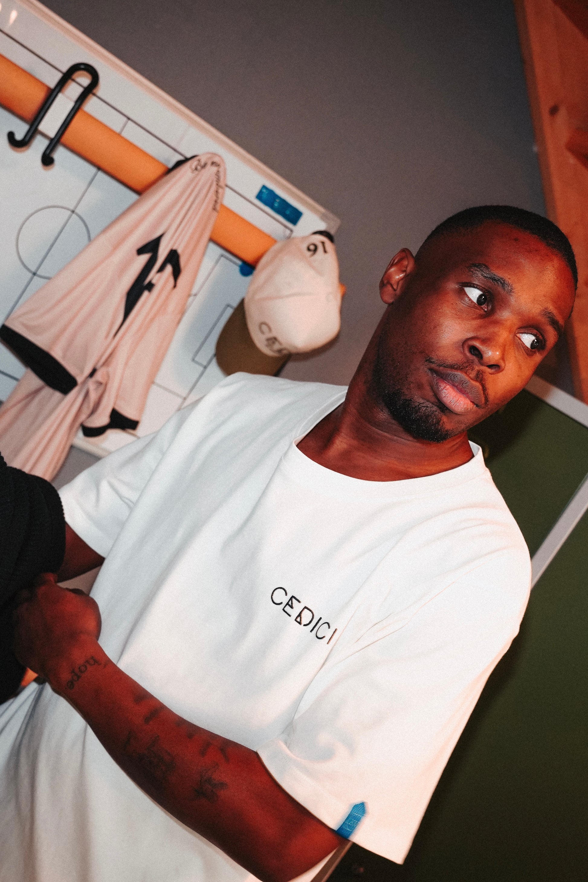 A male model wearing a white CEDICI t-shirt stands in a locker room setting, looking to the side. Behind him, a beige jersey with the number 11 and two caps—one white and one olive—hang from a magnetic whiteboard designed like a football tactics board. His arm features visible tattoos, adding to the casual and athletic atmosphere of the scene. Christian Fassnacht fashion brand.